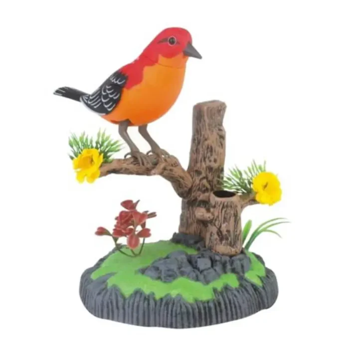 A colorful toy bird perched on a branch, featuring a vibrant red and orange body with black and white wing details. The bird is situated on a realistic-looking branch with green foliage and yellow flowers, all set on a base designed to resemble natural ground with rocks and plants. This toy bird is likely designed to make sounds or be interactive in some way.