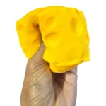 Crunchy yellow cheese block squishy toy with rounded cheese holes, shown in hand and in individual retail packaging with a 12-piece display box, approximately 7 × 7 cm, from SDMAX Toys Wholesaler.